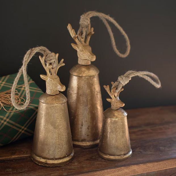Three antiqued gold bells in varying sizes with sculpted deer heads and rope loops, displayed on a wooden surface beside a green plaid gift.