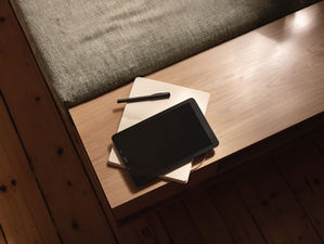 Tablet, pen, and notebooks on a wooden table with a cushion. Warm lighting and wooden floor create a cozy atmosphere. No visible text.