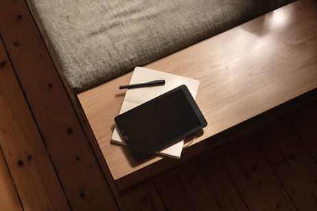 Tablet, pen, and notebooks on a wooden table with a cushion. Warm lighting and wooden floor create a cozy atmosphere. No visible text.