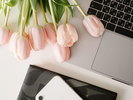 Pink tulips next to a laptop, white phone on a black patterned notebook. Soft lighting and a calm, organized workspace setting.