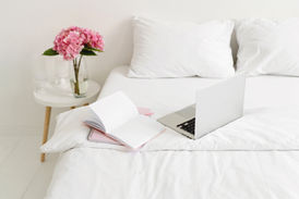Open laptop and notebooks on a white bed, pink hydrangeas in a vase on a side table. Bright, calm, and organized workspace.