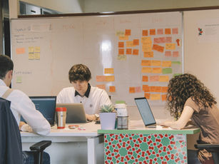 Three people work on laptops at a table in an office. A whiteboard with colourful sticky notes detailing tasks is in the background.
