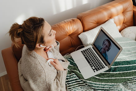A woman wearing earphones takes part in a video call on a laptop while sitting on a sofa.