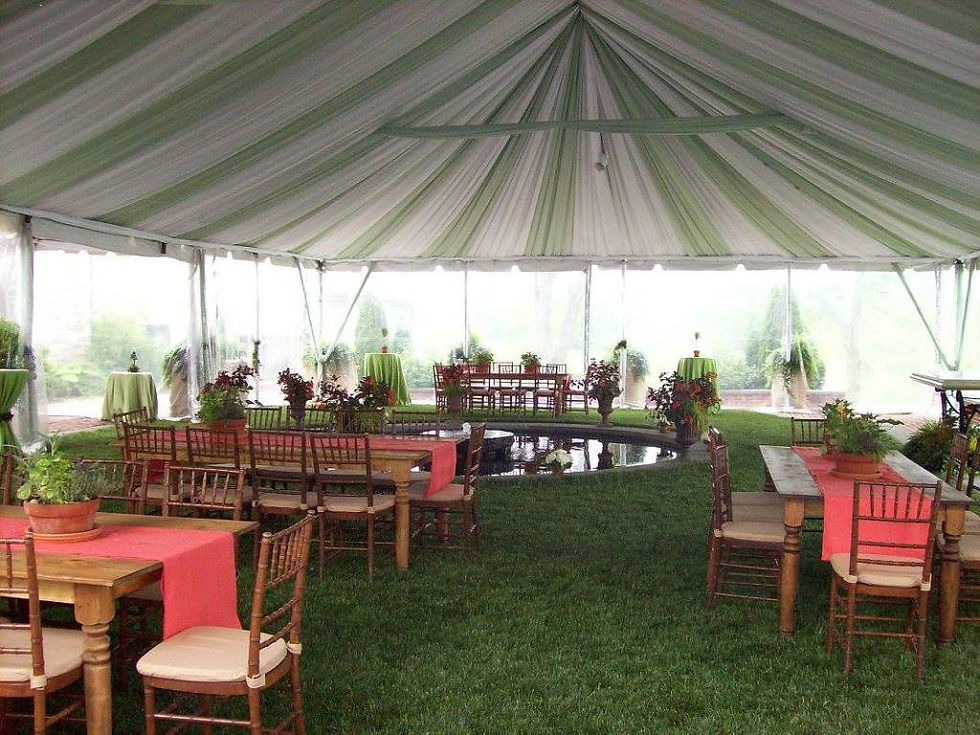Elegant tented venue with wooden tables, red runners, and plants. Green, white draped ceiling, grass flooring, serene outdoor view.