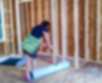 A woman in a black shirt and green bag is measuring wood in a partially constructed room. Rolled materials are on the plywood floor.