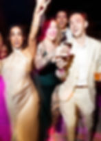 A glamorous group of young people in gold and jewel tone party clothes smile at the photographer while dancing and holding their drinks and phone for pictures