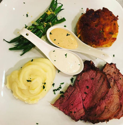 Roast beef slices, mashed potatoes, green beans, and a patty on a plate with two sauces in spoons; garnished with herbs.