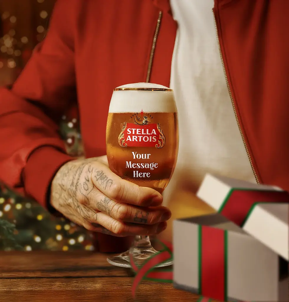 Tattooed hand holding a Stella Artois glass with "Your Message Here", next to gift boxes, against festive background and red jacket.