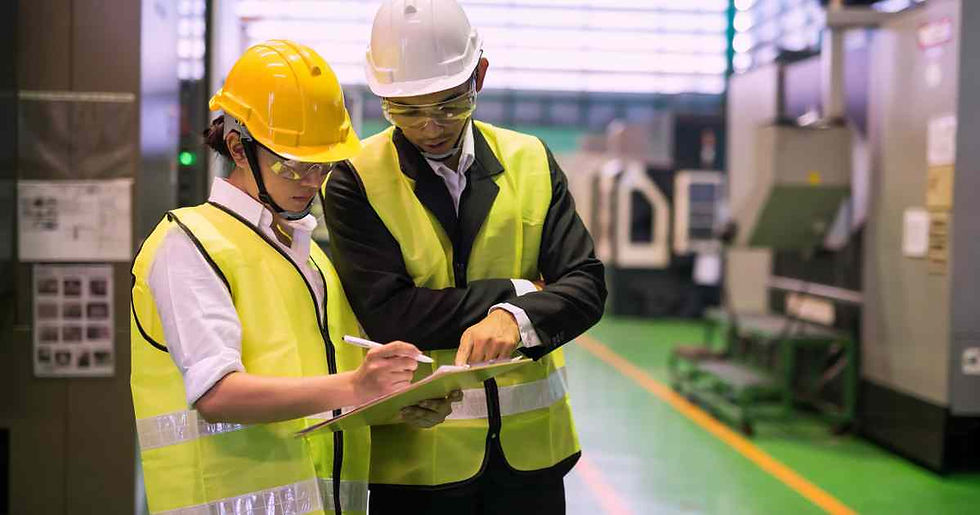 Two people in hard hats and yellow vests review documents on a clipboard in an industrial setting. Machinery is visible in the background.