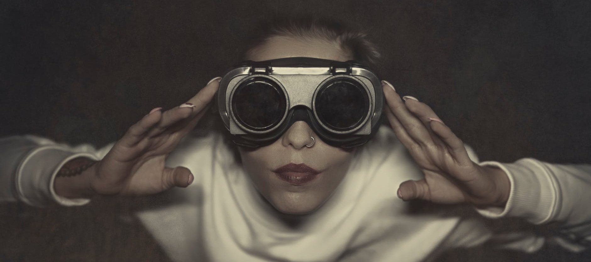 A person in a white hood facing the viewer, both hands raised to a pair of large dark goggles that hide their eyes.