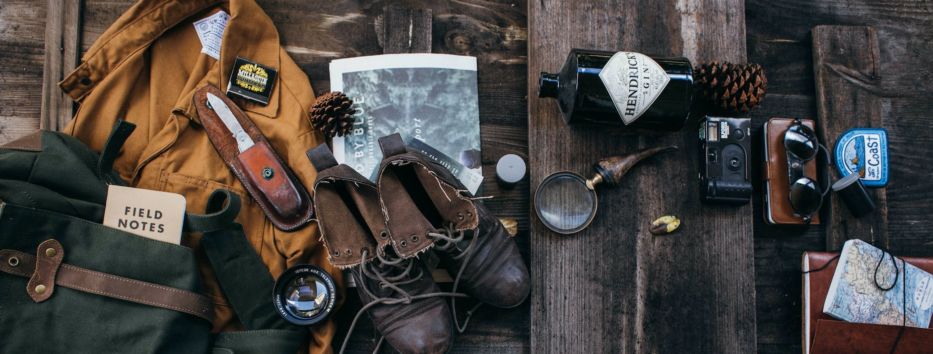 An overhead flat lay of expedition gear on weathered wood: a canvas pack, a field notebook, boots, a jacket, a pocketknife, a camera, and a map.