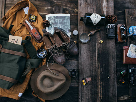 An overhead flat lay of expedition gear on weathered wood: a canvas pack, a field notebook, boots, a jacket, a pocket knife, a camera, and a map.