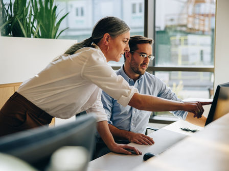 A professional woman and man in a bright office are looking at a computer screen. The woman is pointing to the screen.