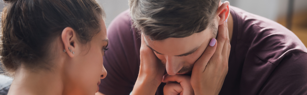 Young woman comforting young man who is depressed. She is putting her hands on his face to offer comfort.