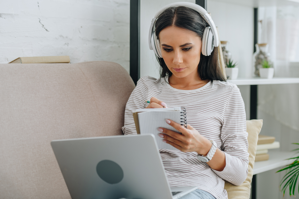 Young woman wearing headphones and taking notes from the laptop computer sitting in her lap