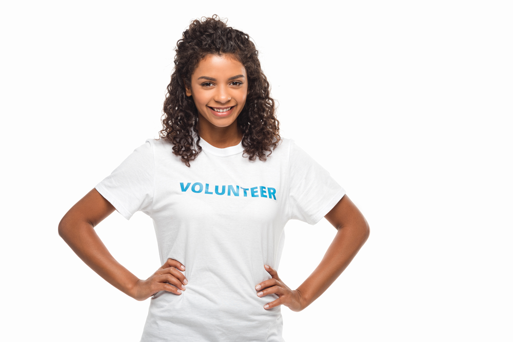 young African-American woman smiling with her hands on her hips. She is wearing a white t-shirt with aqua lettering that says "Volunteer" on it