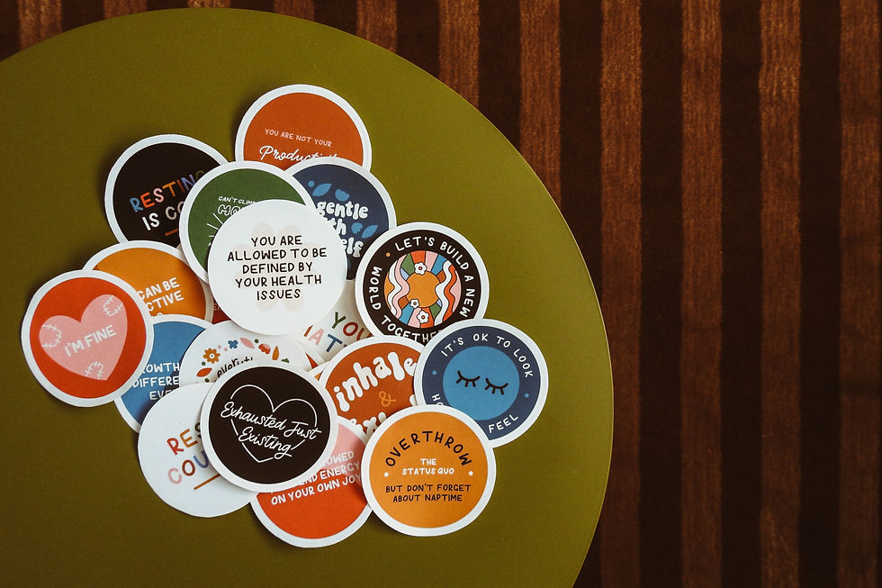 A selection of round stickers displayed on a tabletop. Stickers features phrases such as "exhausted just existing", "I'm fine" and "you are allowed to be defined by your health issues".