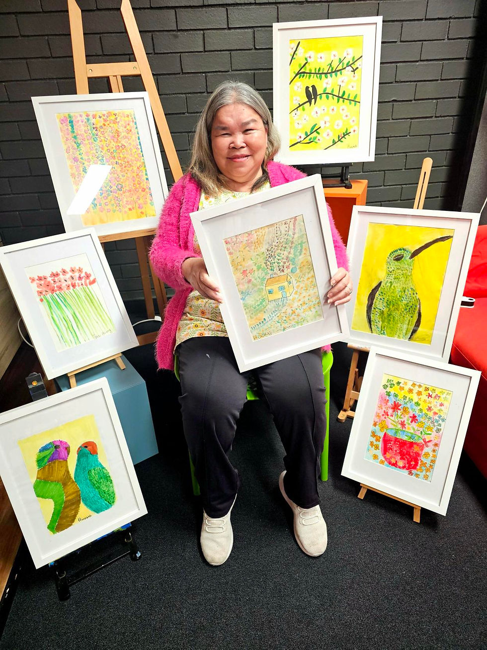 A person is sitting in front of a black wall, holding framed colorful artworks featuring birds and flowers, with similar paintings displayed around.