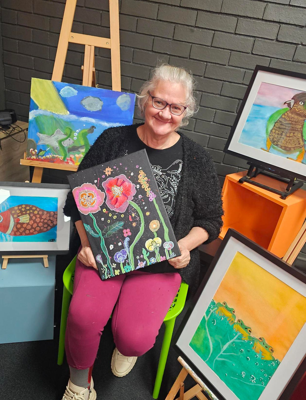 Woman smiling, seated on a green chair, holding colorful floral painting. Surrounded by various vibrant artworks against a black brick wall.