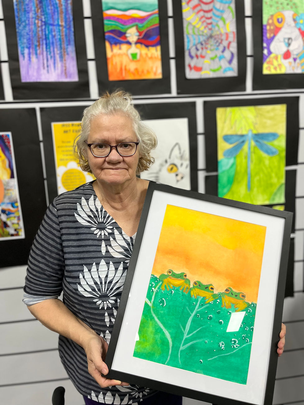 Woman holding a framed painting of three frogs on green leaves against an orange-yellow background. Colorful artwork displayed on wall.