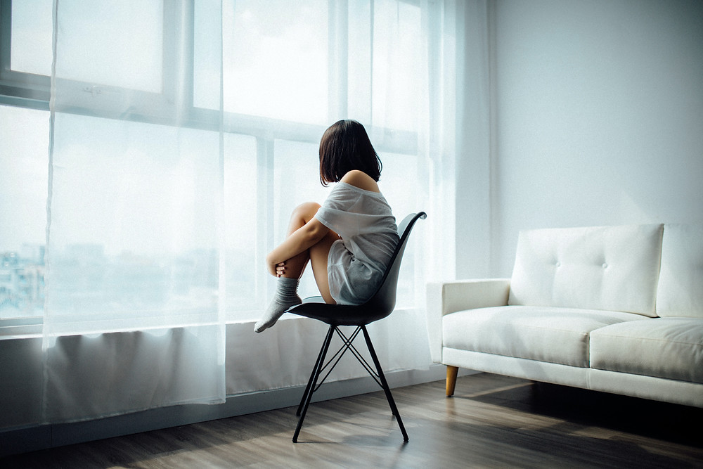 Frau auf Stuhl schaut traurig aus dem Fenster in die Ferne vor Gardine. Frau hat socken und t-Shirt an mit Sofa im Hintergrund