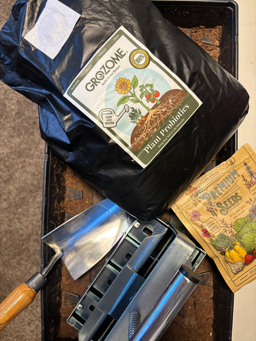 Gardening supplies on soil tray: GroZone plant probiotics bag, Premium Seeds pack, and a metal trowel with a wooden handle, emphasizing home gardening.