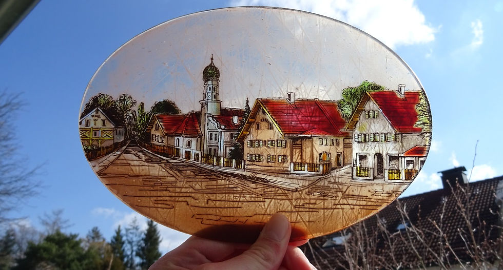 Eine Hand hält ein ovales Glasbild gegen den Himmel, darauf ist der Haderner Dorfkern mit Kirchturm gemalt.