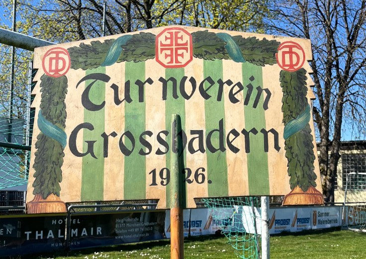Bemalte Holztafel mit der Aufschrift Turnverein Grosshadern 1926, umrahmt von grünen Blattgirlanden.