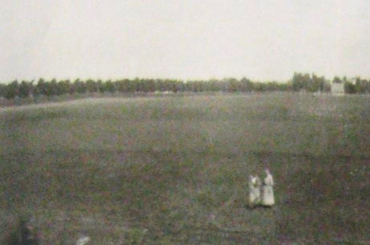 Weite, leere Wiese mit zwei Personen in der Bildmitte, am Horizont eine Baumreihe.