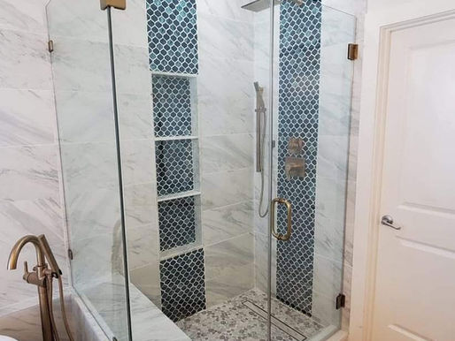 Glass shower with blue mosaic accents and marble tiles in a modern bathroom. Gold fixtures and wooden floor add elegance.