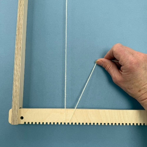 A hand threads white yarn on a wooden weaving loom against a blue background, showcasing a crafting process.