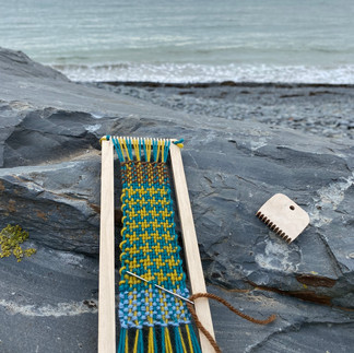 a blue and mustard yellow woven bookmark on a bookmark weaving loom sitting on a rock in front of the sea