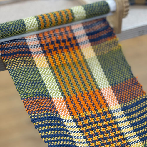 Colorful plaid fabric being woven on a loom with shades of orange, green, blue, and white in a detailed pattern. Indoors, crafting focus.