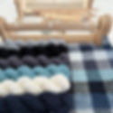 Yarn skeins in blue, white, gray, and black lie near a plaid woven cloth. A wooden weaving loom is in the background on a table.