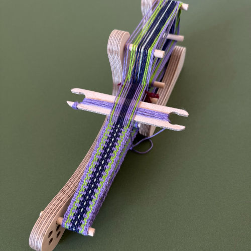 Woven purple, green, and blue strap stretched on a small wooden loom against a plain green background.