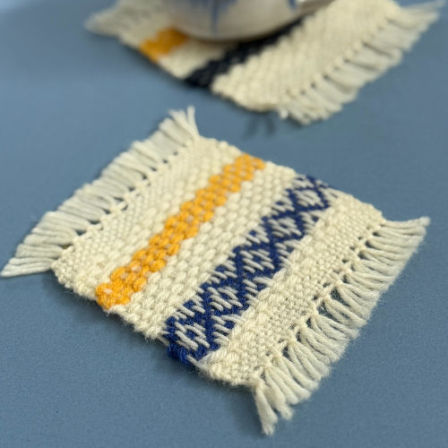 Woven coasters with blue and yellow patterns on a blue surface. Fringed edges and textured design create a cozy, handmade feel.