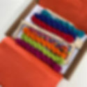 Colorful yarn skeins on a wooden loom frame in an open box with orange tissue paper. Visible text reads "Tabby and Tweed."