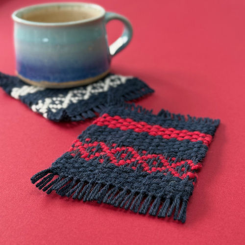 Blue mug on a red surface with a woven coaster featuring red and navy patterns. Another coaster with a cream design is partially visible.