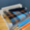 A small loom with blue, black, and orange plaid fabric partially woven. Wooden shuttle and yarn in foreground. Background is plain gray.