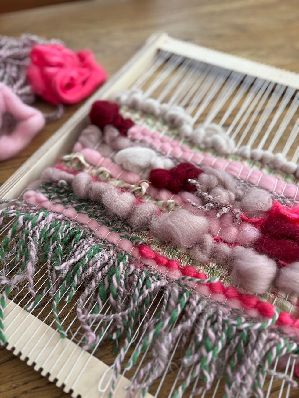 Close-up of a woven textile on a loom with pink, red, and green yarns, set on a wooden surface. Bright, soft textures dominate.
