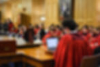 Outward looking view from Parliament Hall. Students dressed in red gowns sitting in the background. Clerk to the house in the foreground working on computer. Debate shield sitting on the table in front of the Clerk.