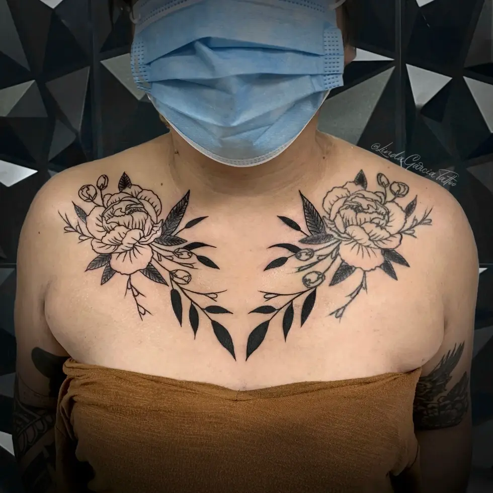 This stunning blackwork collarbone tattoo exudes elegance and grace. Two meticulously rendered peonies form the centerpiece of the design, their delicate petals and buds rendered with impressive detail. The peonies are artfully connected by a symmetrical arrangement of leaves creating an inverted V that visually accentuates the collarbone. The artist's skillful use of negative space enhances the impact of the design, allowing the black ink to pop against the skin. The symmetrical nature of the composition makes it particularly eye-catching and visually appealing. It’s a perfect example of how carefully considered placement can elevate a floral tattoo to a sophisticated piece of body art.