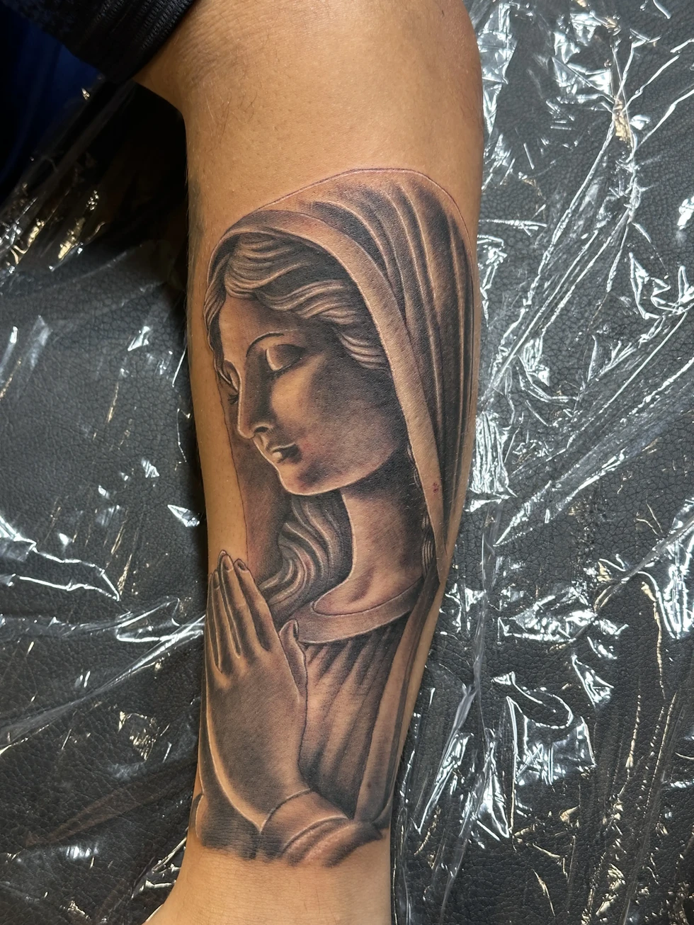 This evocative tattoo, skillfully rendered by the artist Joker, portrays the Virgin Mary in a moment of prayer. The piece is executed in a realistic black and grey style, with meticulous attention to detail. The shading and detail used to depict Mary's serene expression and the folds of her veil are particularly noteworthy. The tattoo captures a sense of peace and reverence, making it a powerful piece of religious art. The placement on the arm allows for the design to be easily visible, a constant reminder of faith. This tattoo is a beautiful example of how religious iconography can be translated into a timeless piece of body art.