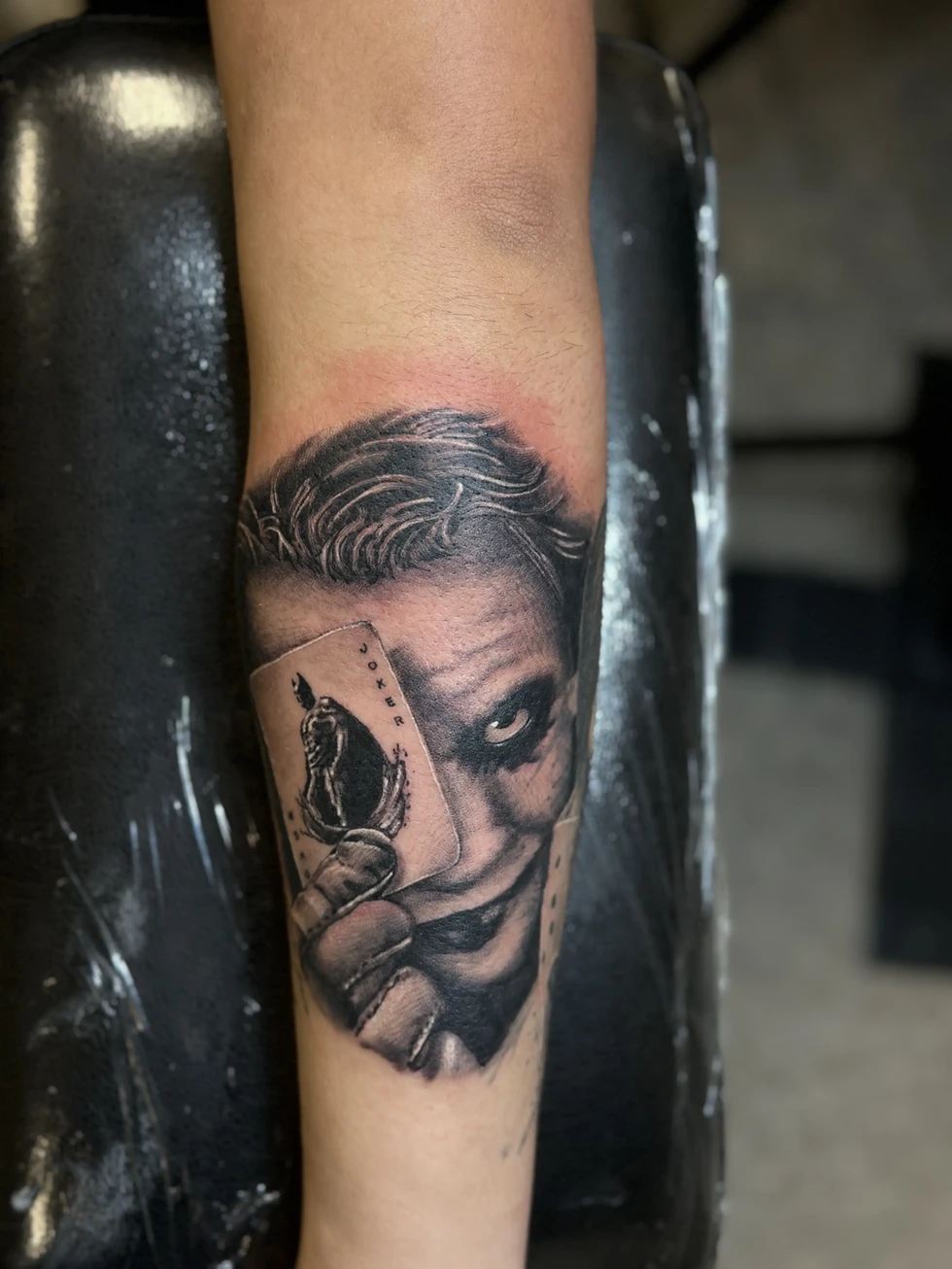 This striking tattoo by Joker Ink is a realistic portrayal of the iconic villain, the Joker. Rendered in black and grey, the tattoo captures the Joker's menacing grin and intense gaze. He's depicted holding a Joker playing card, a subtle detail that adds to the overall impact of the design. The artist's skill in shading and detail is evident in the texture of the Joker's hair and the folds of his skin. The placement on the forearm makes this a bold statement piece. This tattoo is a testament to Joker Ink's ability to create captivating and detailed portraits. The fact that this is a longtime client speaks to the artist's consistent quality and ability to build rapport.