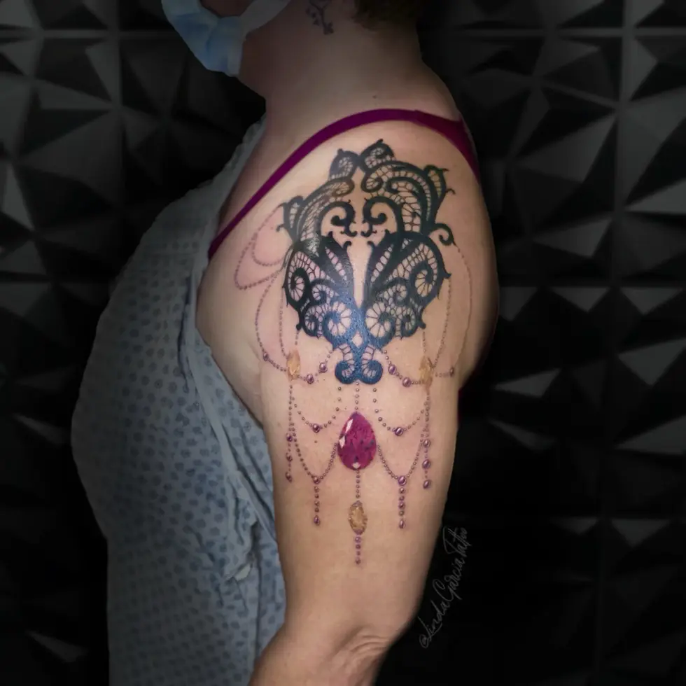 This exquisite shoulder tattoo is a stunning example of how body art can mimic luxurious textiles and jewelry. The tattoo is designed to resemble intricate black lace, with delicate details that create a realistic and visually captivating effect. Adding to the opulence, the lace is adorned with strands of beads in shades of purple and amber, culminating in a striking, centrally placed pink gemstone. The tattoo drapes elegantly over the shoulder, creating a unique and feminine piece. The combination of the lace-like blackwork and the colorful jewels results in a tattoo that is both sophisticated and eye-catching.