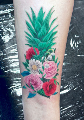 Color forearm tattoo of a pineapple formed from pink, red and pale flowers, topped with a crown of pointed green leaves.