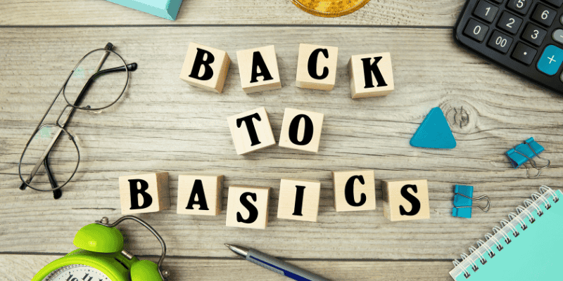 Wooden blocks on a desk spelling out 'Back to Basics', surrounded by everyday office items including glasses, an alarm clock, calculator, notebook, pen, and clips, representing a return to fundamental principles.