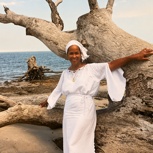 Smiling woman in white dress and headwrap stands by giant driftwood on a beach with the sea behind her.
