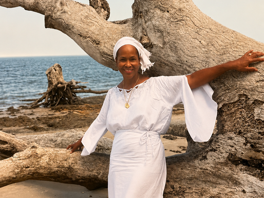 Smiling woman in white dress and headwrap stands by giant driftwood on a beach with the sea behind her.