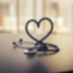 A stethoscope forms a heart shape on a smooth, dark surface. Blurred background with warm tones. Represents care and compassion.